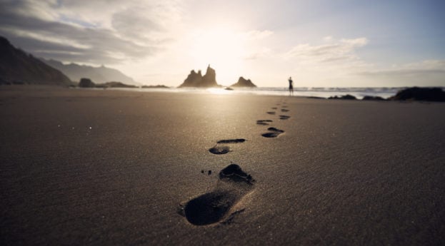 Perspectiva de baixo para cima de pegadas de pessoa na areia caminhando em direção ao mar, alusivo à felicidade