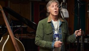 Paul McCartney, líder da lista de turnês mais lucrativas do mundo, de jaqueta verde, em estúdio escuto e microfone, cantando