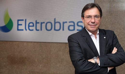Wilson Ferreira Júnior, de terno preto e camisa branca e óculos, sorrindo de boca fechada, em frente a um backdrop da Eletrobras