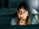 Mulher de óculos de armação de plástico transparente e franja olhando para tela de computador, alusivo ao pensamento nos investimentos por perfil de investidor