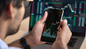 Close de homem de costas, usando óculos, ampliando tela de celular com gráficos de mercado financeiro, alusivo às ações para investir em setembro