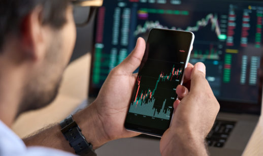 Close de homem de costas, usando óculos, ampliando tela de celular com gráficos de mercado financeiro, alusivo às ações para investir em setembro