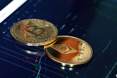 Conceito de duas criptomoedas representando Bitcoin e Ethereum, sobre tela de tablet com gráficos de mercado financeiro
